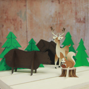 forest paper animals in a paper campsite. pictured is a bear, deer, and squirrel in front of trees.